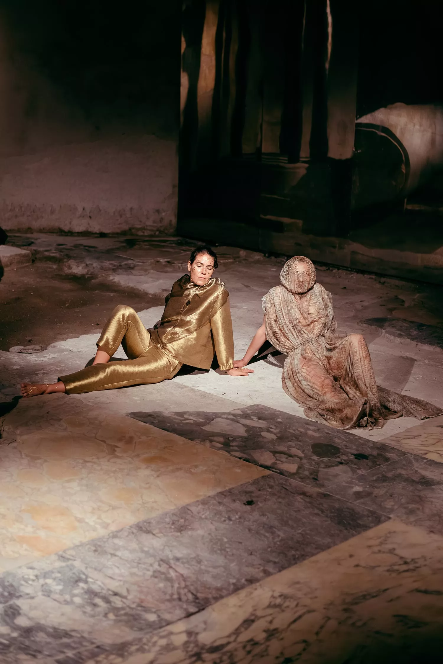 Gaia Fugazza - Gaia Fugazza, 2024, Farewell to Known Skies. Two performers sit at the centre of a beige tiled floor: one wears a gold suit, while the other is covered in netting. A dark, indistinct background surrounds them.