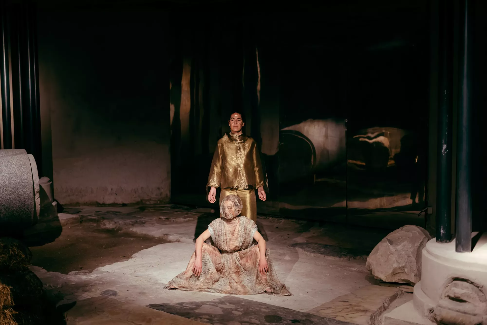 Gaia Fugazza - Gaia Fugazza, 2024, Farewell to Known Skies. Two performers occupy a dark, atmospheric space surrounded by large columns and stones. One sits wearing a long netted dress and face covering, while another stands behind them in a metallic gold outfit.